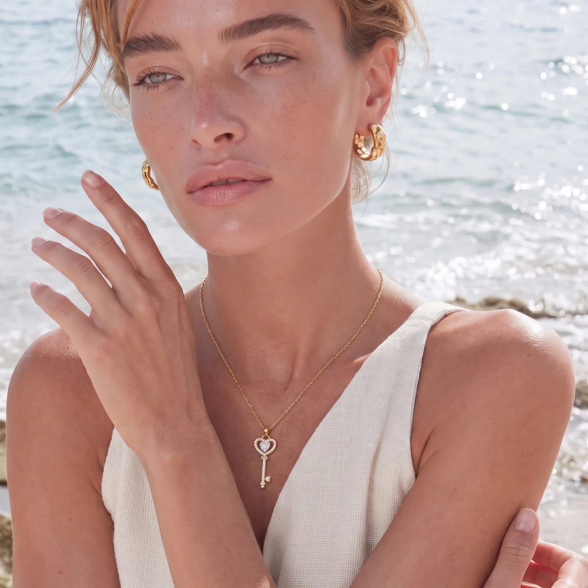 Woman by the sea wearing a gold heart key pendant and gold hoop earrings