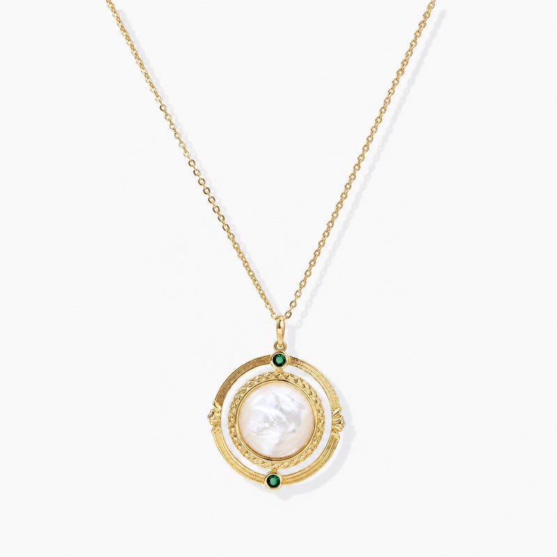 Gold plated necklace with a round white MOP pendant, textured gold ring, and two green zirconia stones on a cream background.