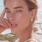 Gold-plated flower stud earrings with textured petals and centered freshwater pearl on beige background