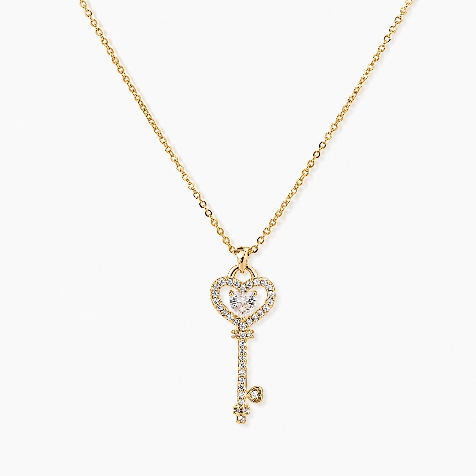 Delicate gold key pendant with heart-shaped bow and crystal pavé detailing on fine chain against cream background.
