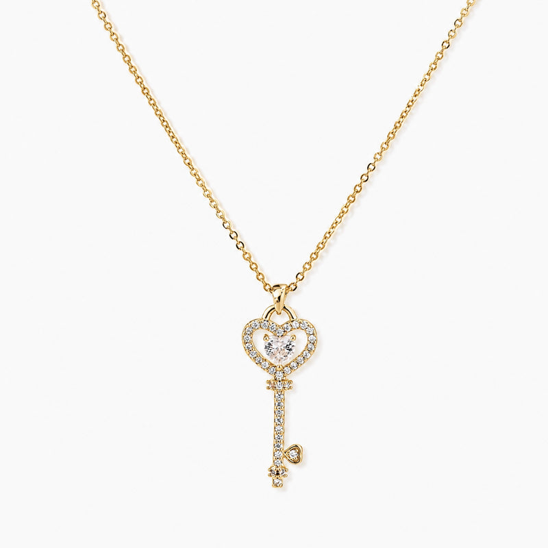 Delicate gold key pendant with heart-shaped bow and crystal pavé detailing on fine chain against cream background.