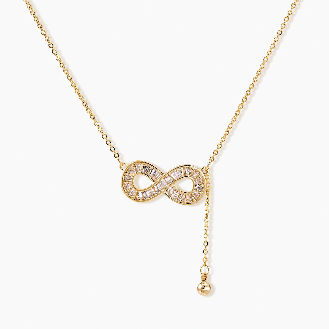 Gold infinity pendant with baguette crystals on adjustable chain against cream background.
