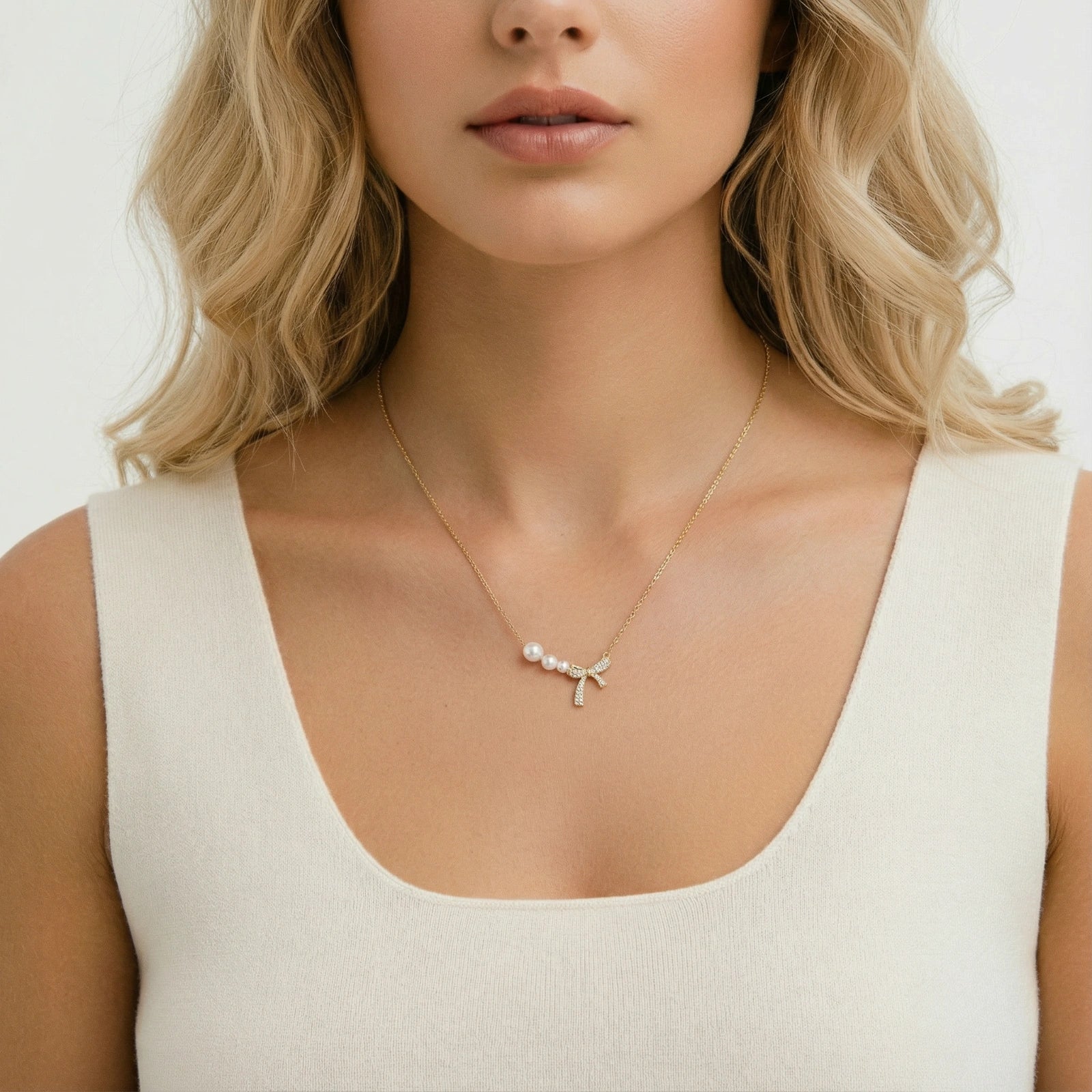 Delicate gold necklace with three white pearls and crystal-embellished bow pendant on cream background