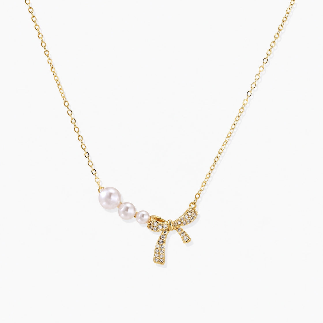 Delicate gold necklace with three white pearls and crystal-embellished bow pendant on cream background