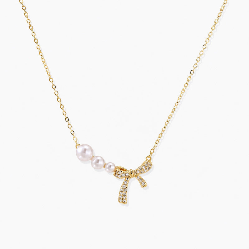 Delicate gold necklace with three white pearls and crystal-embellished bow pendant on cream background