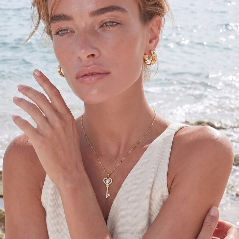 Woman by the sea wearing a gold heart key pendant and gold hoop earrings